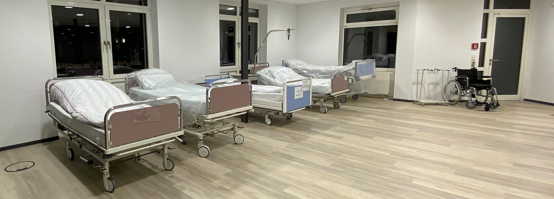 Mehrere Krankenhausbetten und ein Rollstuhl sind abgebildet: In den Demoräumen können die Auszubildenden praktische Fertigkeiten in der Pflege, Pflegetechniken und Aufgaben wie Injektionen, Infusionen oder Lagerungen üben.
