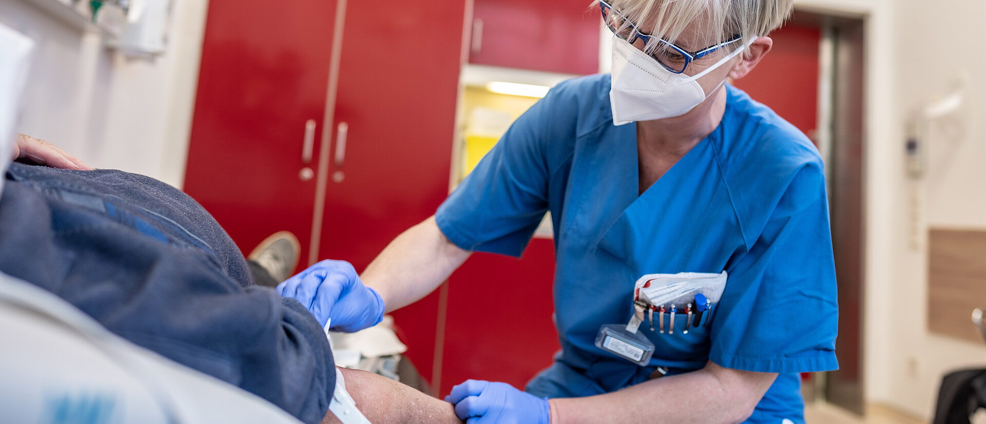 Pflegekraft der Zentralen Notaufnahme kniet neben Patienten und nimmt Blut ab. Dill-Kliniken, Dillenburg.