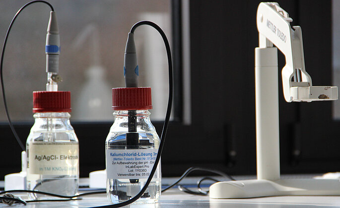 Zwei Glasflaschen vor Abzugshaube mit Elektroden. 